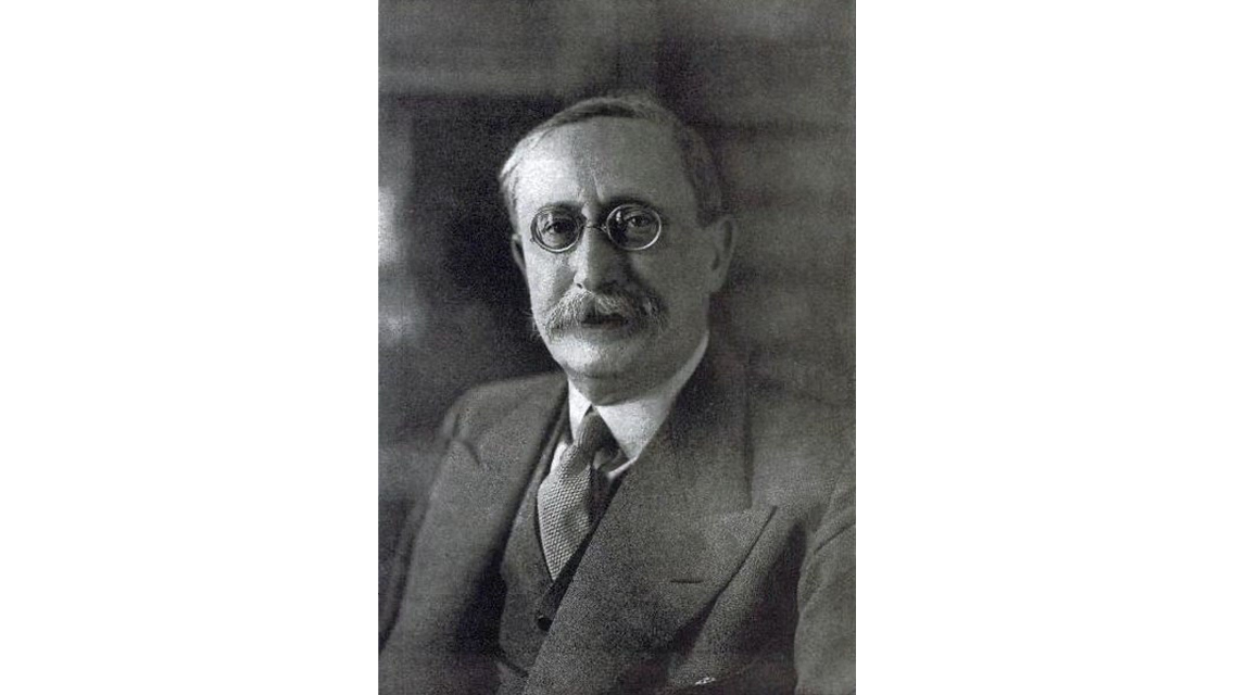 Black-and-white portrait photograph of an older man with round wire-rimmed glasses and a moustache, wearing a suit and tie. 
